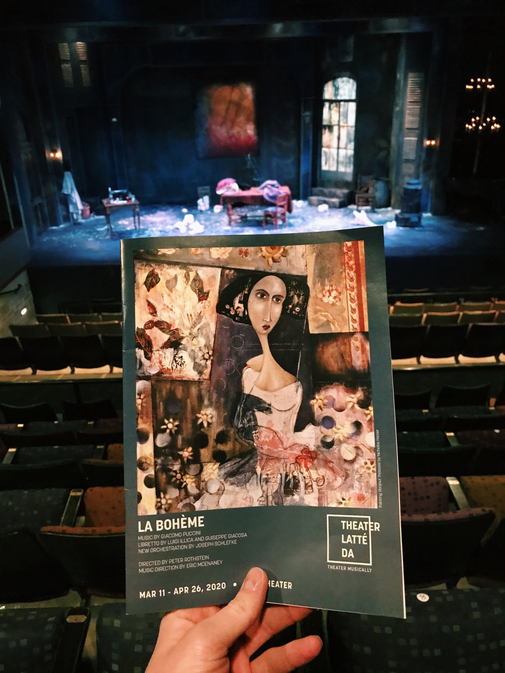 A hand holding a theater program in front of chairs and the stage