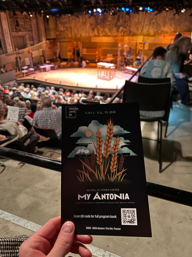 A hand holding a theater program that reads "My Antonia" with theater seats and a stage in the background