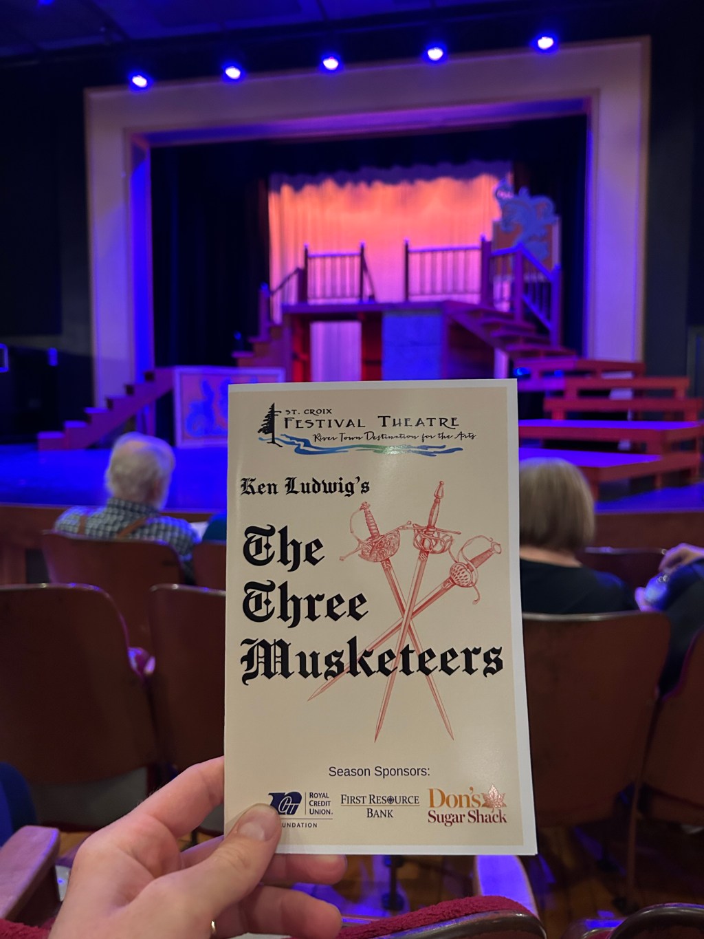 A hand holding up the program for "The Three Musketeers" at St. Croix Festival Theatre with the stage in the background