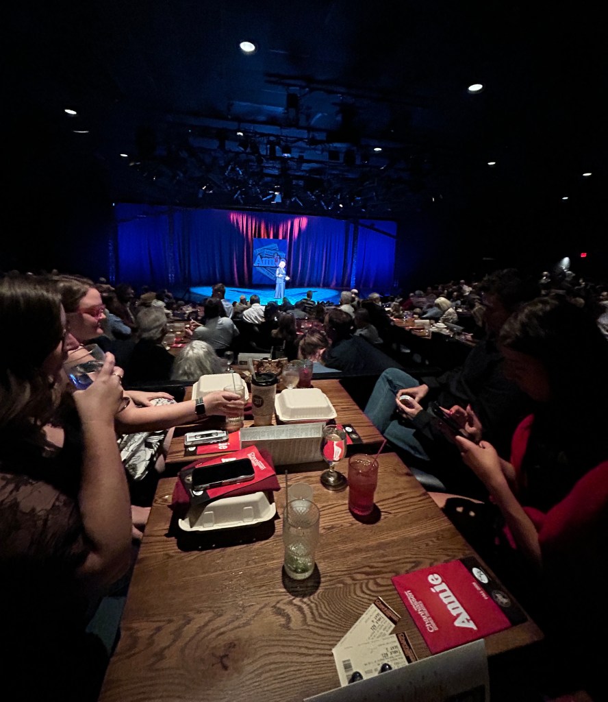 Three women and one man sit at tables with a stage in the background bathed in blue light that says "Annie"